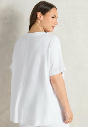 Vrouw met lang bruin haar, gekleed in een loszittende witte top met korte mouwen, kijkt over haar schouder tegen een egale lichte achtergrond.