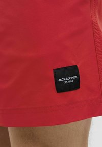 Červené plavecké šortky z hladkého materiálu, s černou visačkou s bílým textem, na které je napsáno "JACK & JONES EST. 1990." Šití viditelné podél okrajů.