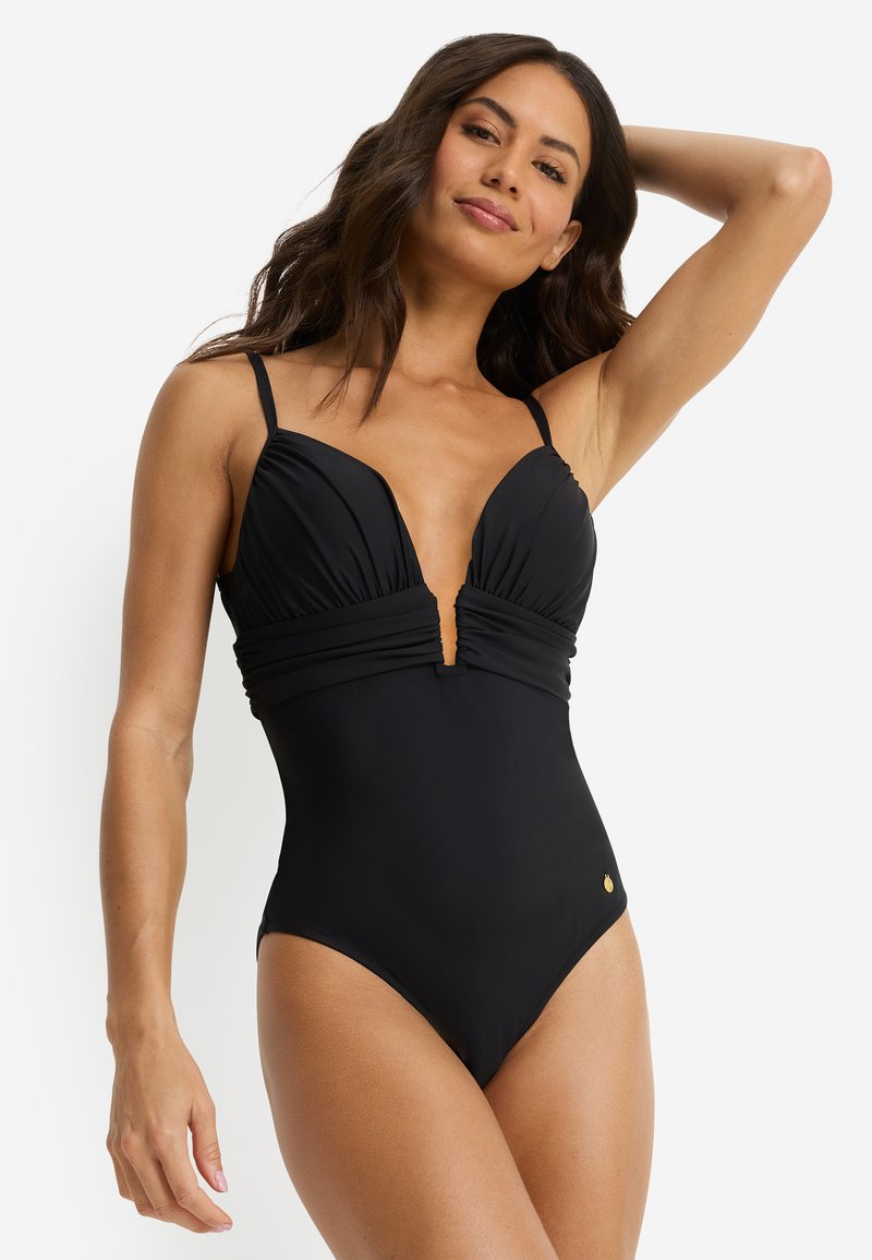 Μαύρο ολόσωμο μαγιό με βαθιά V λαιμόκοψη, ραφές που τονίζουν το στήθος, ρυθμιζόμενους ιμάντες και μικρό χρυσό λογότυπο. Λεία υφή υφάσματος.