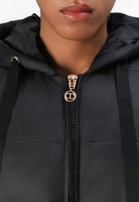 Musta toppatakki hupulla, jossa on kultainen vetoketju ja logotyyppitys. Sisältää mustat kangasnauhat ja sileän, kiiltävän pinnan.