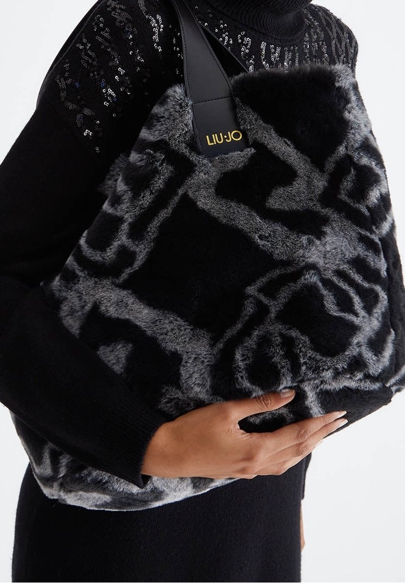 Personne portant un pull noir à sequins tenant un grand sac à main Liu Jo en fausse fourrure noire et grise avec une sangle en cuir noir et un logo doré.