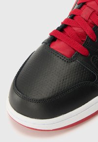 Fekete bőr sportcipő texturált felülettel, piros fűzőkkel és fehér középtalppal, piros talppal. Lekerekített orrforma.