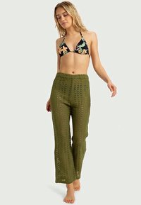 Pantalones de ganchillo en verde oliva, diseño de pierna ancha, ligeros y con textura estampada, combinados con un top de bikini triangular negro con flores.