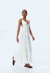 Witte mouwloze maxi-jurk met een diepe V-hals, gelaagde stoffen en een zwierige zoom. Gecombineerd met bruin geweven instapsandalen.