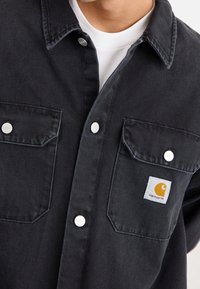 Black denim shirt with two chest pockets and silver snap buttons. Features a yellow Carhartt logo patch on the left pocket. Classic design.