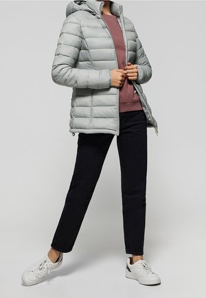 Personne portant une veste matelassée gris clair, un sweat-shirt bordeaux, un pantalon noir et des baskets blanches, debout avec un pied légèrement levé.