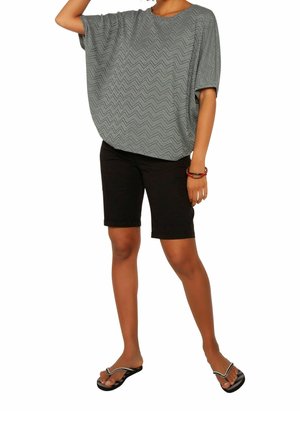 Personne portant un haut ample gris à motif zigzag, un short noir arrivant aux genoux, des tongs noires et des bracelets rouges et noirs à un poignet.