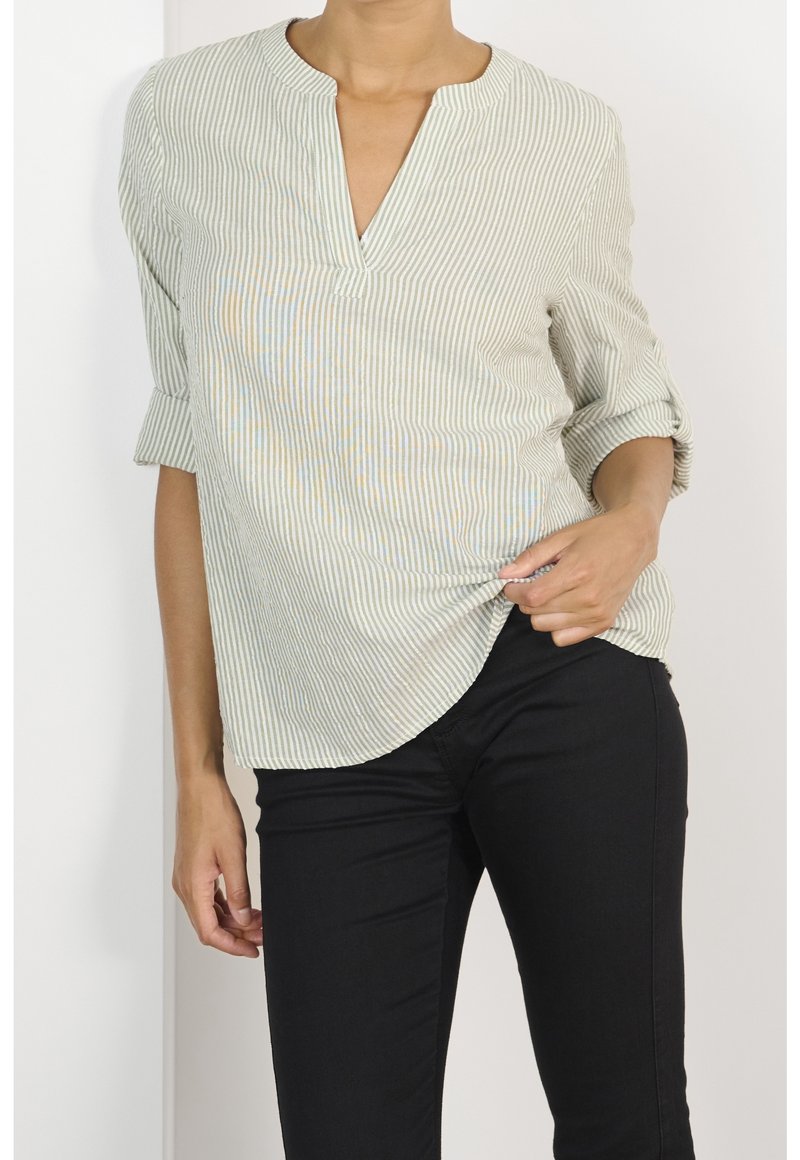 Light green striped blouse with a half-placket, roll-up sleeves, and curved hem. Worn with black pants, featuring a smooth texture.