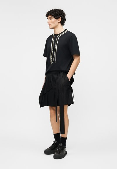 Mladý muž s kudrnatými vlasy, který má na sobě černé tričko s krátkými rukávy a vzorovanými šňůrkami, černé zavinovací šortky s dlouhými pásky, černé ponožky a černé tenisky.