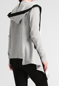 Sudadera gris con un acento negro, diseñada con un dobladillo trasero largo y un acabado texturizado. Cuenta con capucha y mangas largas.