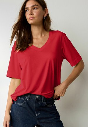 Barna hajú nő, ezüst karika fülbevalóval, piros V-nyakú pólóval és sötétkék farmernadrággal, aki úgy pózol, hogy az egyik kezét a csípőjén tartja.