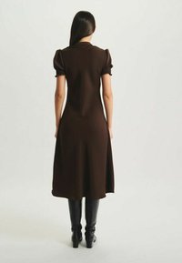 Robe marron mi-longue avec des manches bouffantes, tissu lisse et jupe évasée. Associée à des bottes noires au genou, vue de l'arrière.