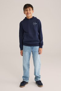 Námořnická mikina s kapucí, přední kapsou a logem na prsou, v kombinaci se světle modrými džíny a tmavými teniskami.