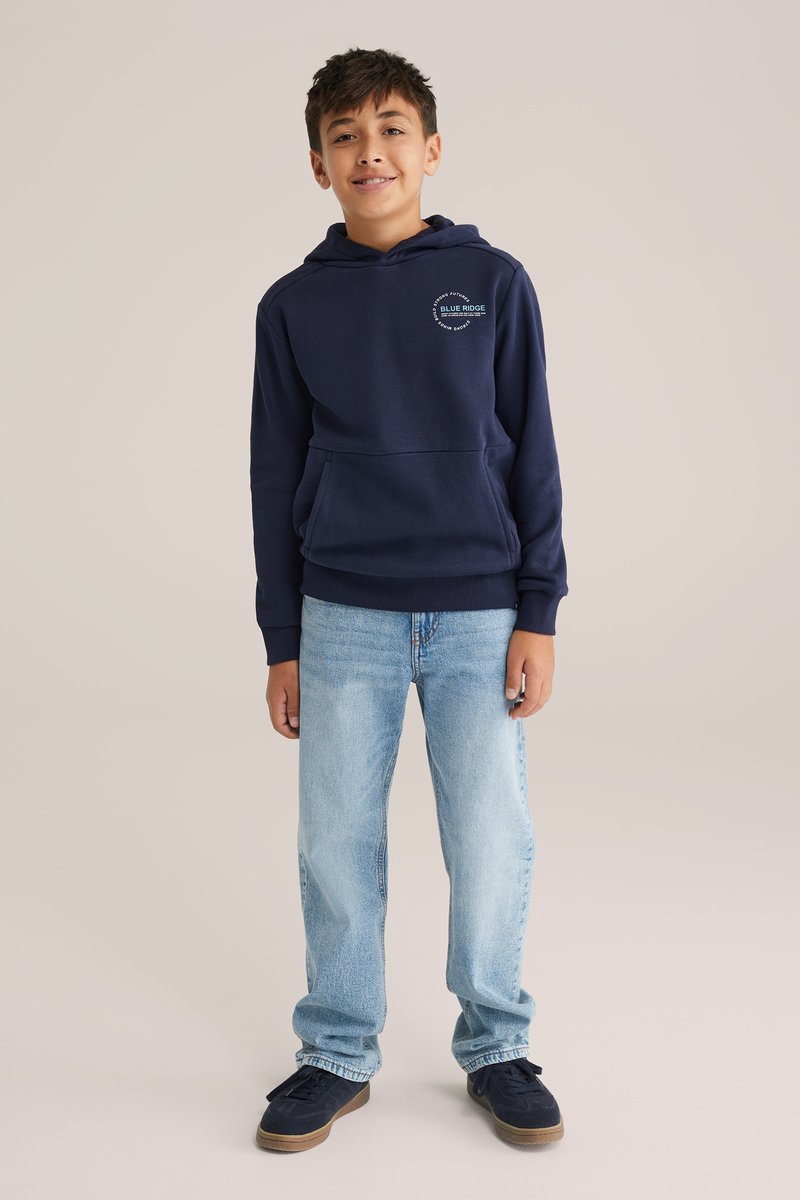 Námořnická mikina s kapucí, přední kapsou a logem na prsou, v kombinaci se světle modrými džíny a tmavými teniskami.