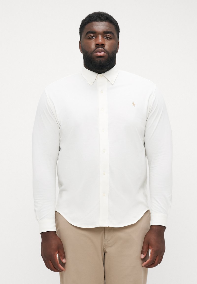 Chemise blanche à manches longues avec un col boutonné, présentant un logo discret sur la poitrine gauche. Texture lisse, ourlet droit et tissu léger.