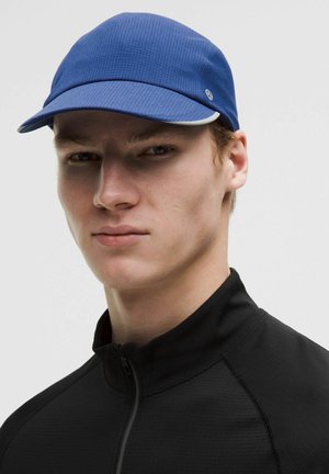 Young man wearing a blue sports cap and black zip-up top, facing forward with a neutral expression against a plain light background.