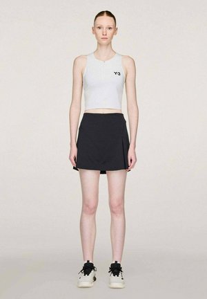 Jonge vrouw staande en recht vooruit kijkend, met een witte Y-3 mouwloze cropped top, een zwarte minirok en witte sneakers met zwarte veters.