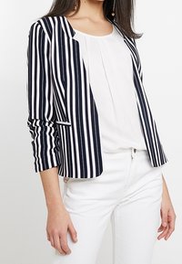 Blazer rayé marine et blanc avec une coupe ajustée, revers crantés et manches trois-quarts. Porté sur un haut blanc et associé à un pantalon blanc.