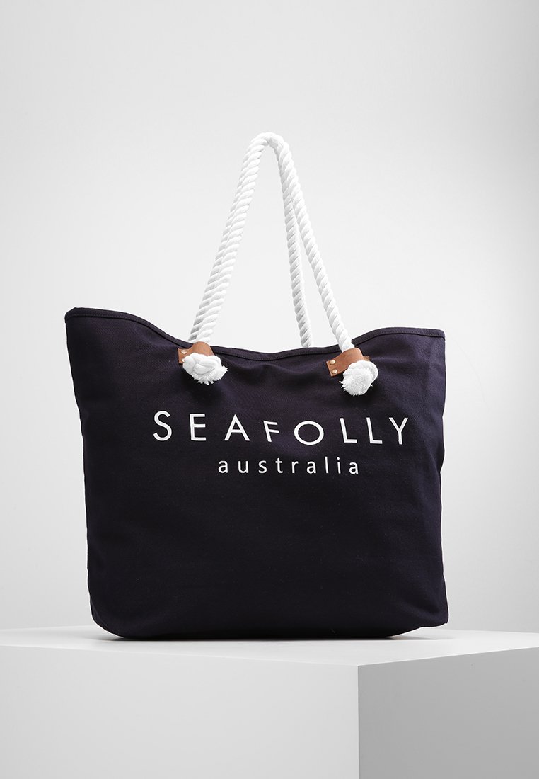 Mörkblå canvas tote-väska med vita repetaxlar, som har en logotyp och texten "SEAFOLLY australia" i vit tryck. Mjuk textur, rymlig design.
