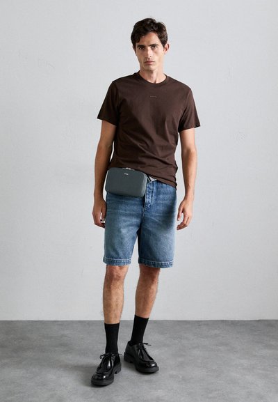Maglietta girocollo marrone, pantaloni corti di jeans, scarpe nere e calze. Una borsa grigia con zip è indossata in vita, caratterizzata da un design minimal.