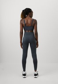 Žena s pletenom kosom u tamnosivoj sportskom brusnom i legginsima, s bijelim čarapama i crnim tenisicama, stoji okrenuta leđima prema jednostavnoj pozadini.