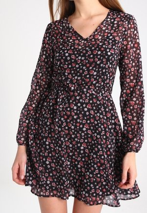 Zwarte lange mouw jurk met klein rood-wit bloemenpatroon, V-hals, getailleerde taille en licht uitlopende rok, gedragen door een persoon.