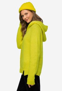 Světle zelený pletený svetr s kapucí, s ribkovanými manžetami a lemem. V páru s odpovídající pletenou čepicí, vystaveno na světlém pozadí.