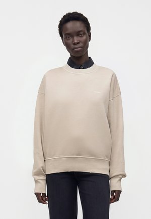 Woman wearing beige oversized sweatshirt over black collared shirt and dark pants, standing against plain light background.
