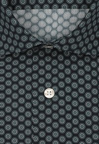 Camicia con bottoni su sfondo scuro, caratterizzata da cerchi concentrici in teal e bianco, con un bottone bianco sulla patta. Tessuto morbido.