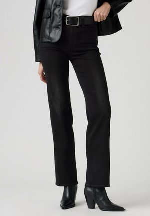 Persona che indossa jeans neri a gamba dritta, cintura nera, top bianco, giacca di pelle nera e stivali neri con tacco, in piedi davanti a uno sfondo neutro.