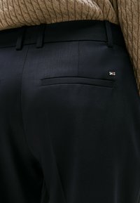 Zwarte op maat gemaakte broek met een zachte textuur, zijzak en een klein logo-accent op de achterzak.