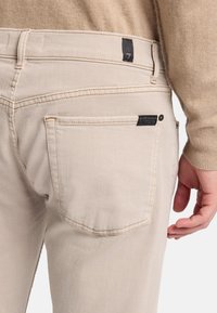 Jeans di denim beige con una vestibilità slim, dotati di tasche posteriori visibili, cuciture a contrasto e un'etichetta nera del marchio sulla vita.