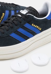 Černé semišové tenisky s modrými pruhy a zlatým textem "Gazelle", s tlustou bílou podrážkou. Součástí jsou dvě sady bílých tkaniček.