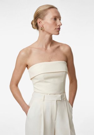 Kobieta o blond włosach związanych w niski kok, ubrana w kremowy top bez ramiączek i pasujące do niego spodnie z wysokim stanem, z rękami w kieszeniach, zwrócona bokiem.