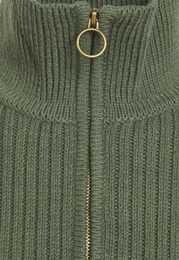 Zielony sweter z prążkowanego materiału z wysokim kołnierzem, z złotym zamkiem i wyraźną fakturą. Ozdobiony pionowymi wzorami dzianiny.