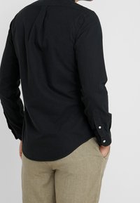 Muž v černé košili s dlouhým rukávem a béžových kalhotách, se založenýma rukama v kapsách, pohled zezadu na jednobarevném pozadí.