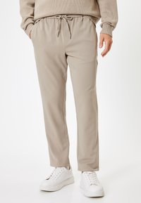 Beige joggerbukser med elastisk talje og snor. Glat tekstur, tætsiddende design, sidelommer, parret med hvide sneakers.