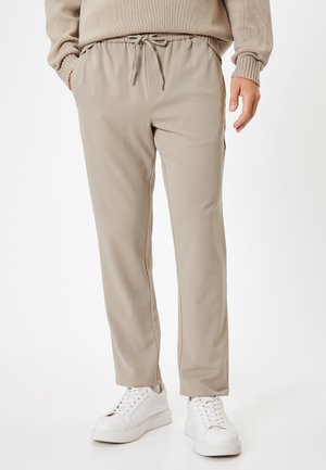 Beige joggerbukser med elastisk talje og snor. Glat tekstur, tætsiddende design, sidelommer, parret med hvide sneakers.
