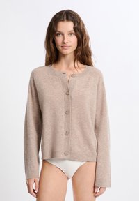 Frau mit welligem braunem Haar, die eine beigefarbene, geknöpfte Strickjacke und weiße Bikinihosen vor einem einfarbigen weißen Hintergrund trägt.