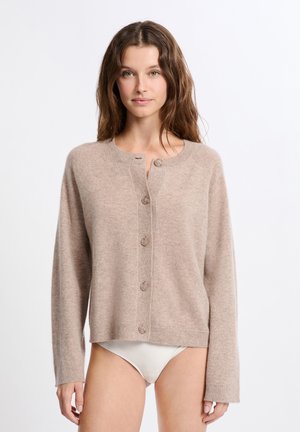 Vrouw met golvend bruin haar, gekleed in een beige knoopcardigan en witte bikini-broekjes tegen een simpele witte achtergrond.