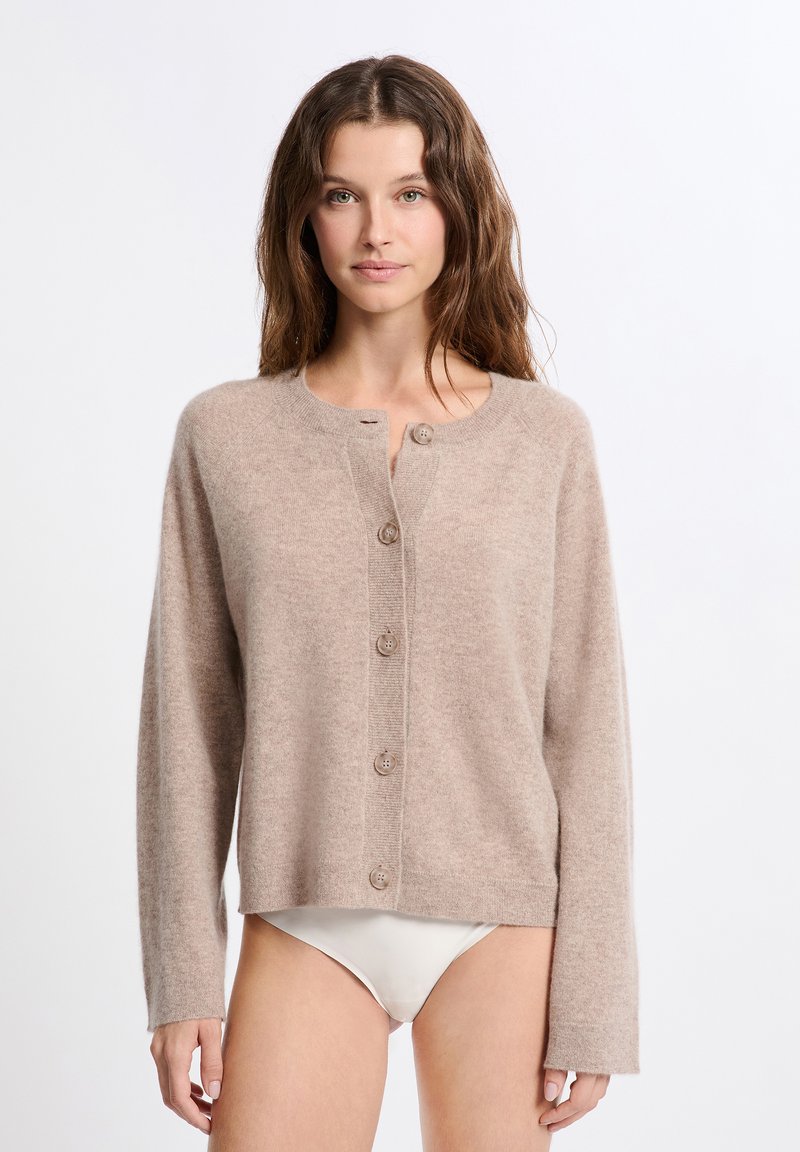 Frau mit welligem braunem Haar, die eine beigefarbene, geknöpfte Strickjacke und weiße Bikinihosen vor einem einfarbigen weißen Hintergrund trägt.