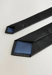 Noeud papillon noir avec une surface texturée et des extrémités pointues. La couche intérieure présente un tissu bleu à pois blancs. Forme géométrique, design épuré.