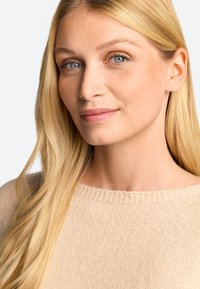 Svetlo bež pulover z rebrastim ovratnikom, ki ga nosi oseba z dolgimi, ravnimi blond lasmi in jasnimi modrimi očmi. Discreten, naraven makeup.