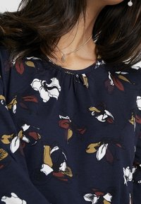 Blouse marine présentant un motif floral en blanc, bordeaux et moutarde, avec un col rond et un détail de tissu froncé aux épaules.