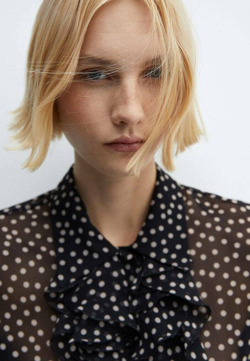 Schwarze Bluse mit weißen Punkten, Rüschensaum und transparentem Stoff; enthält helle beige Punkte, elegantes Design und weiche Textur.