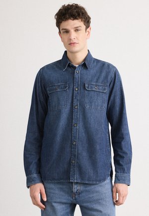 Sötétkék denim ing, hegyes gallérral, két mellzsebbel, gombos előlappal és hosszú ujjakra szabva. Sima textúra varrási részletekkel.