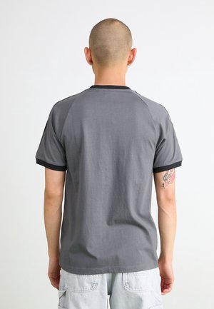 Mand med barberet hoved og tatovering på venstre arm iført grå T-shirt med sort kant og lyse cargoshorts, set bagfra mod hvid baggrund.
