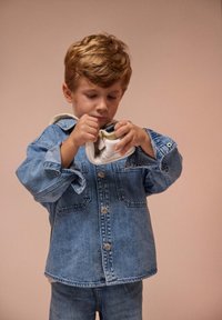Ensemble en jean de couleur bleu clair, composé d'une veste et d'un pantalon, avec des boutons argentés et une capuche. Enfant tenant une chaussure blanche, concentré sur les détails des lacets.