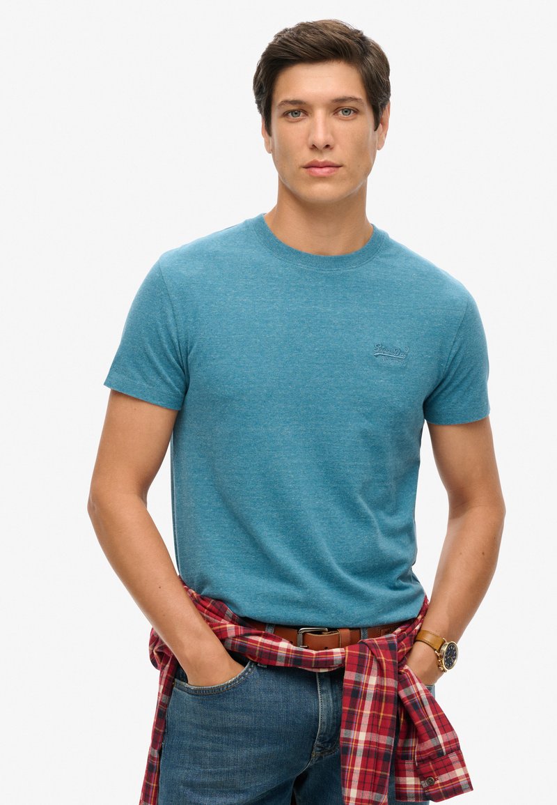 Blauwe T-shirt met korte mouwen en ronde hals, gemaakt van gestructureerde stof. Model draagt een rood en zwart geruit shirt geknoopt om de taille en denim jeans.