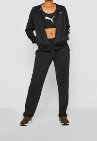 Conjunto atlético negro que incluye una sudadera con cremallera y una camiseta de tirantes corta. Los pantalones son de corte holgado. Todas las prendas están adornadas con el logotipo blanco de Puma.